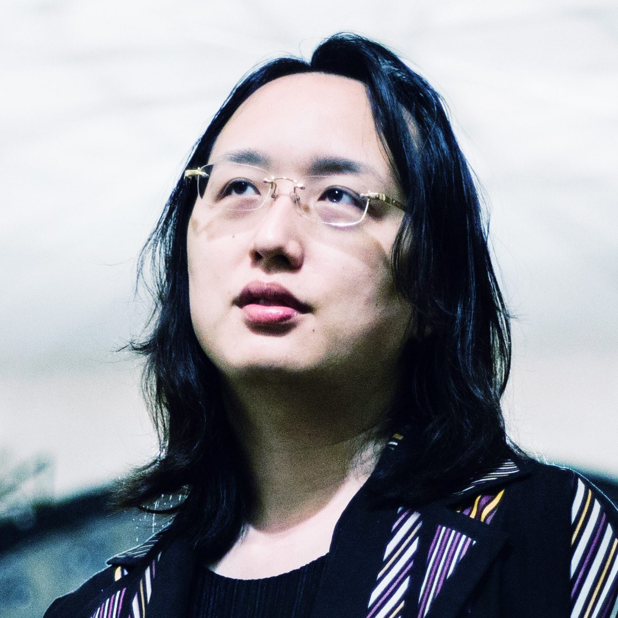 Audrey Tang gazing upward, wearing rimless glasses and a dark striped blazer, in a close-up portrait with soft lighting
